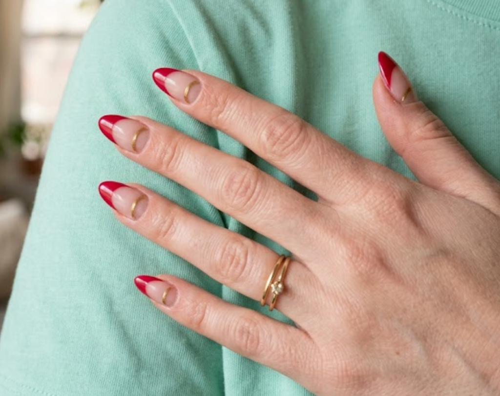 In this close-up photo, Chinese New Year nails featuring almond negative space half-moon tips with crimson tips and gold dividers are shown on a real hand wearing a mint shirt.