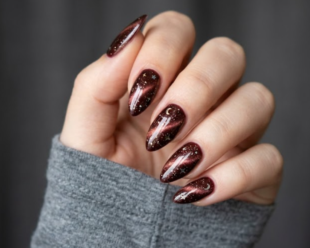 In this close-up photo, Chinese New Year nails featuring stiletto deep burgundy cat-eye tips with scattered starry shimmer effects are shown on a real hand wearing a soft gray shirt.