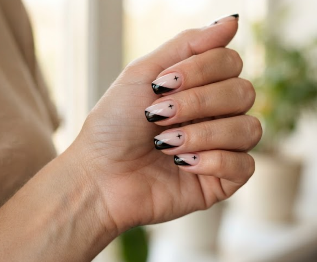 In this image, a woman's full hand displays nude and white nails with black diagonal swipes and star stamps, wearing a beige shirt.