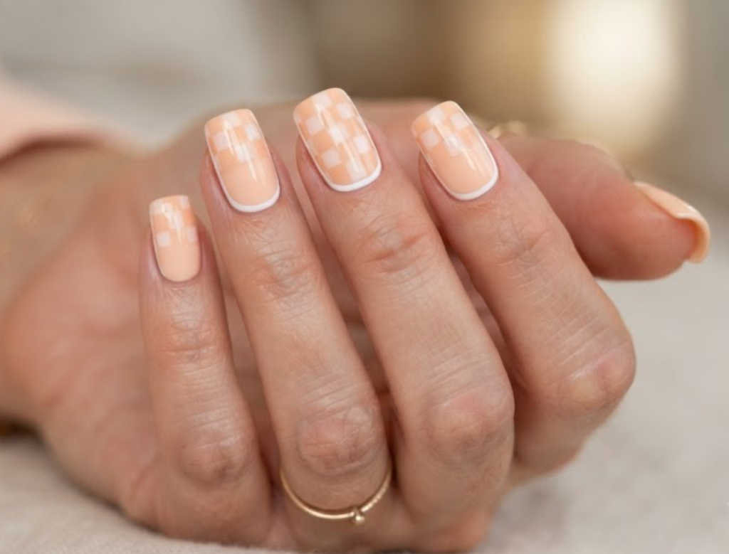 In this image, a woman's full hand features peach nails with upper-half checkerboard grid patterns and thin borders, wearing a peach shirt.