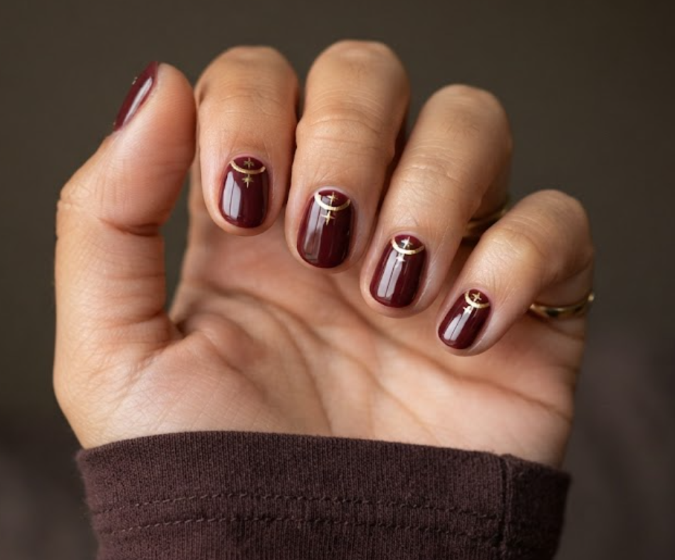 In this image, a woman's full hand displays maroon nails with gold cuticle crescent lines and starburst accents, wearing a brown shirt.