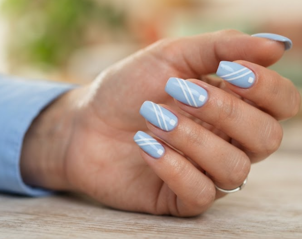 In this image, a woman's full hand highlights pastel blue nails with three diagonal slashes and a white square, wearing a sky shirt.