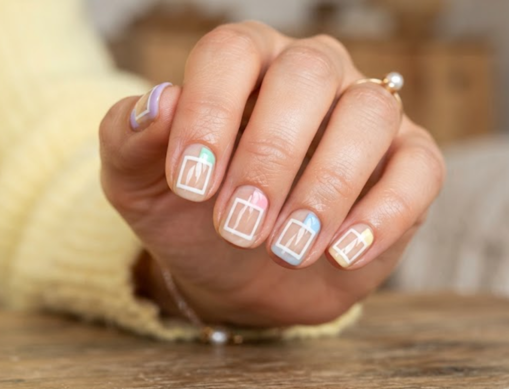 In this image, a woman's full hand highlights transparent nails with white square outlines and pastel blocks, wearing a yellow shirt.