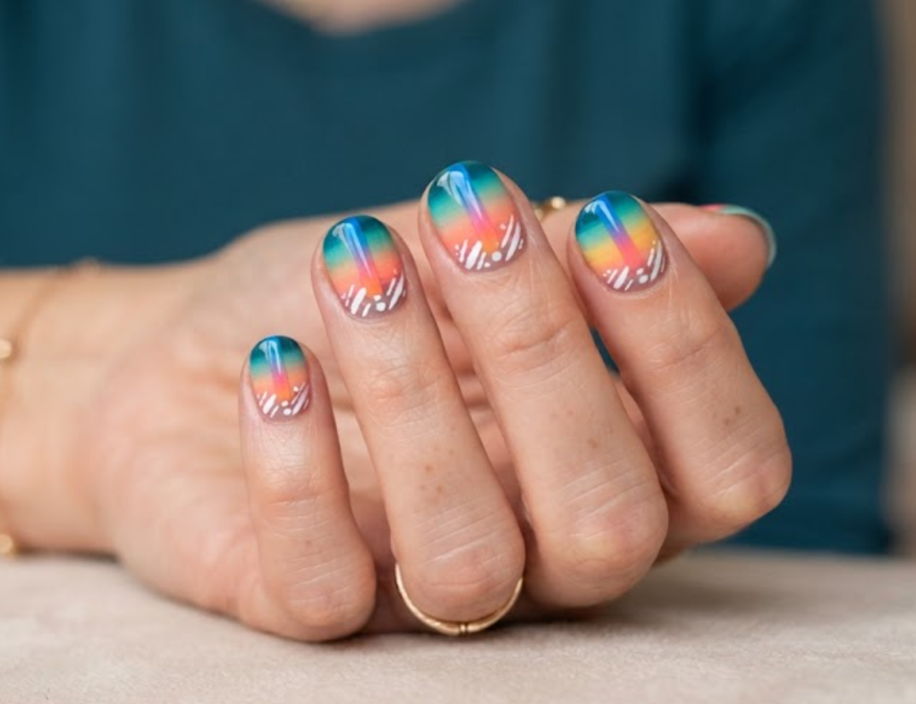 In this image, a woman's full hand shows multi-color gradient nails with central vertical ombre stripes and white slashes, wearing a teal shirt.