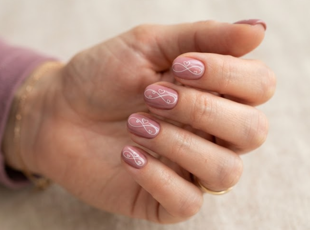 In this image, a woman's full hand features dusty rose nails with continuous infinity loop lines and outline hearts, wearing a mauve shirt.age