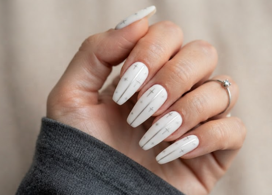 In this image, a woman's full hand features matte white nails with silver cross patterns and sparkle asterisks, wearing a charcoal shirt.