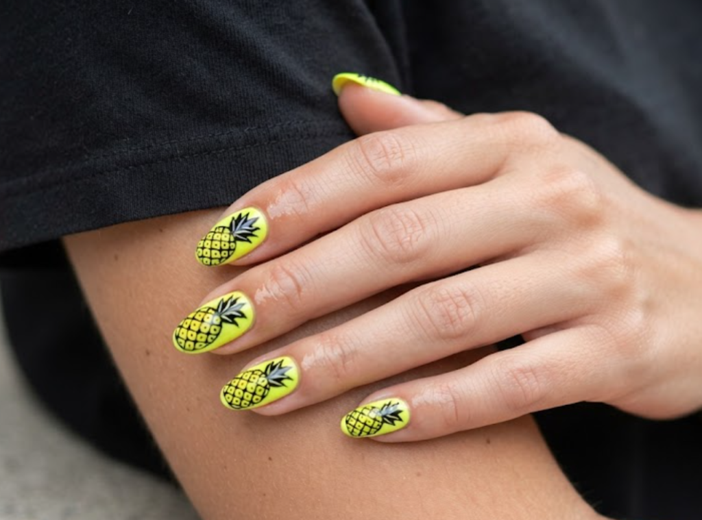 imain this image, a hand in a black shirt features neon yellow and black contrast pineapple nails.