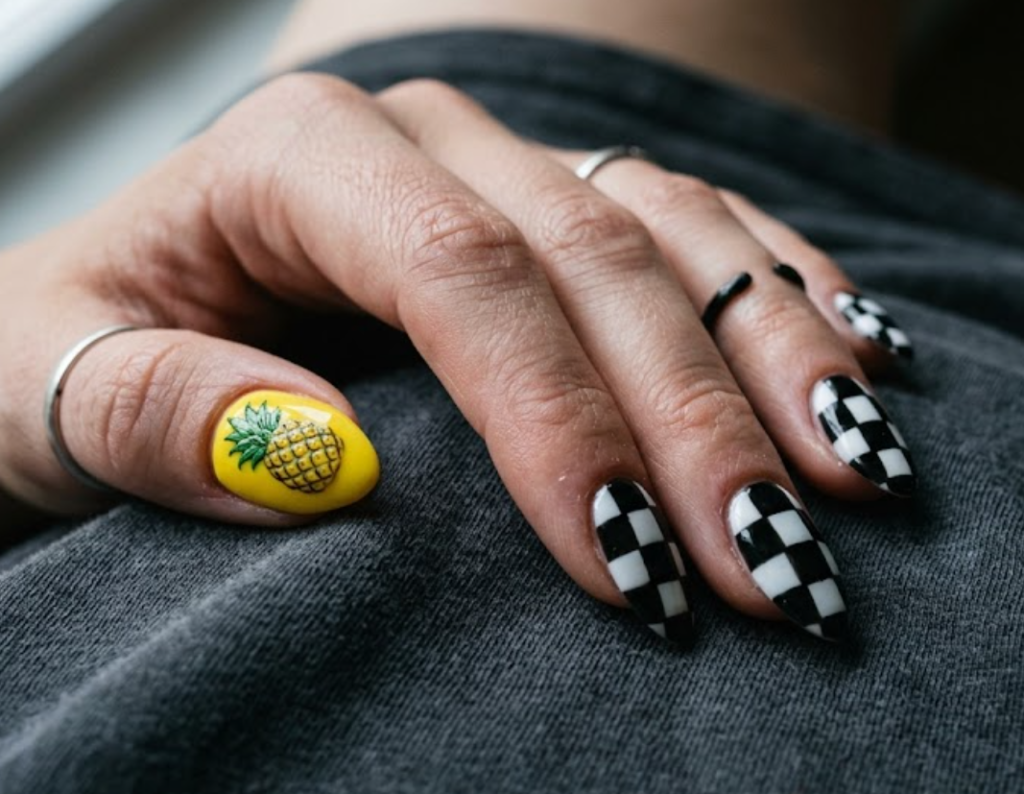 in this image, a hand in a grey shirt showcases black and white checkerboard nails with a yellow pineapple.