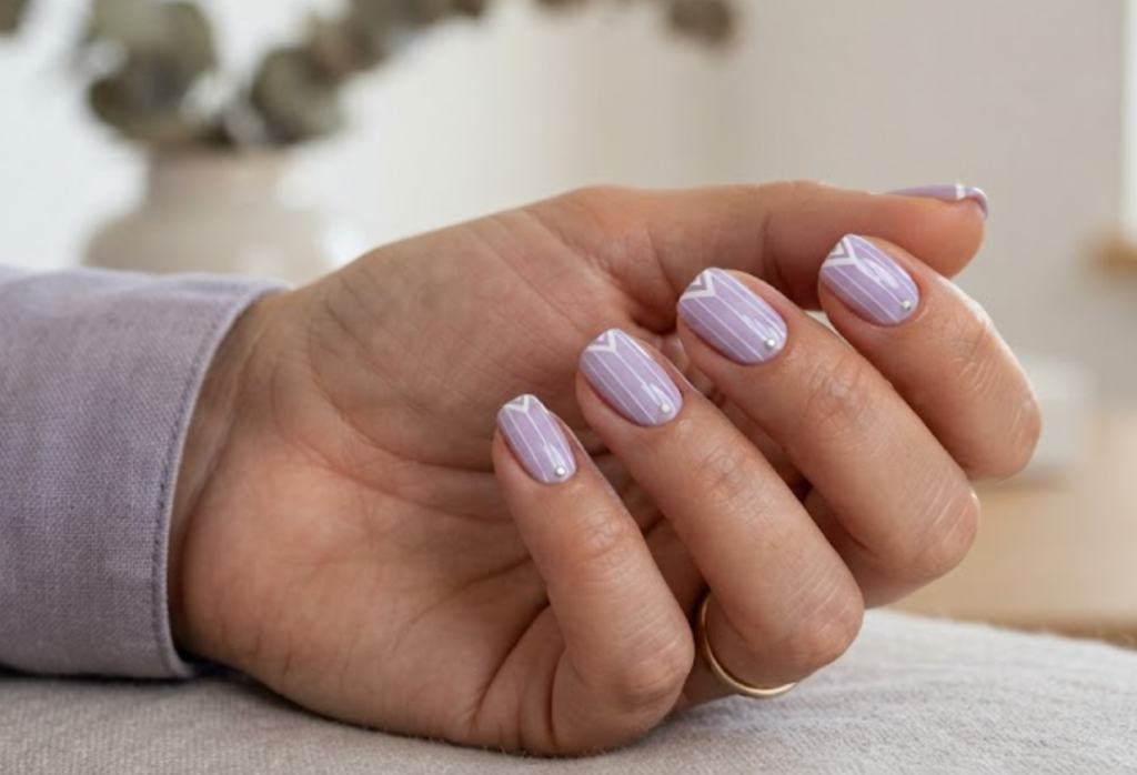 In this image, a woman's full hand showcases lavender nails with vertical lines, metallic dots, and chevron tips, wearing a grey shirt.